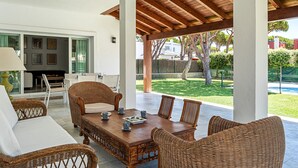 A covered patio with wicker furniture, a wooden table, and a view of a swimming pool and greenery.