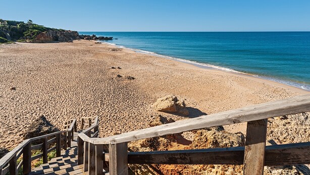 A beach with a wooden staircase leading down to the sand, a clear blue sky, and calm sea.