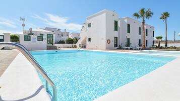 A swimming pool with a metal ladder, white buildings with green shutters, and palm trees.