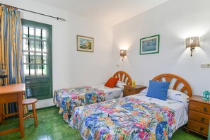 A bedroom with two beds, a wooden desk, a chair, a green door, and wall-mounted lamps.