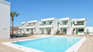 Una piscina con plataforma de concreto blanco, una palmera y un edificio con balcones y una escalera.