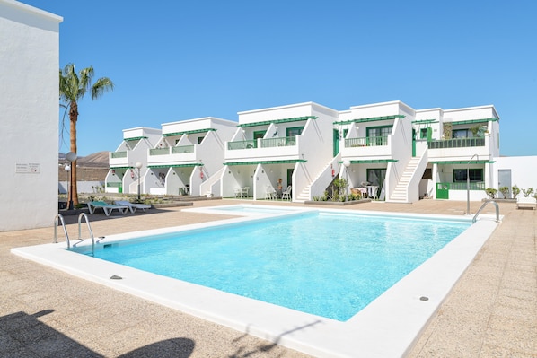 A swimming pool with a white pool deck, a palm tree, and a building with balconies and a staircase.