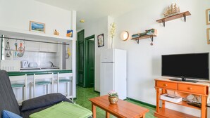 Un espacio habitable compacto con kitchenette, un sofá con cojines, una mesa de madera, un televisor sobre un mueble bajo y un estante pequeño con objetos decorativos.