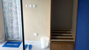 Una habitación con un armario de madera, un tapete azul con la palabra “WELCOME”, una cesta blanca y zapatillas en el suelo.