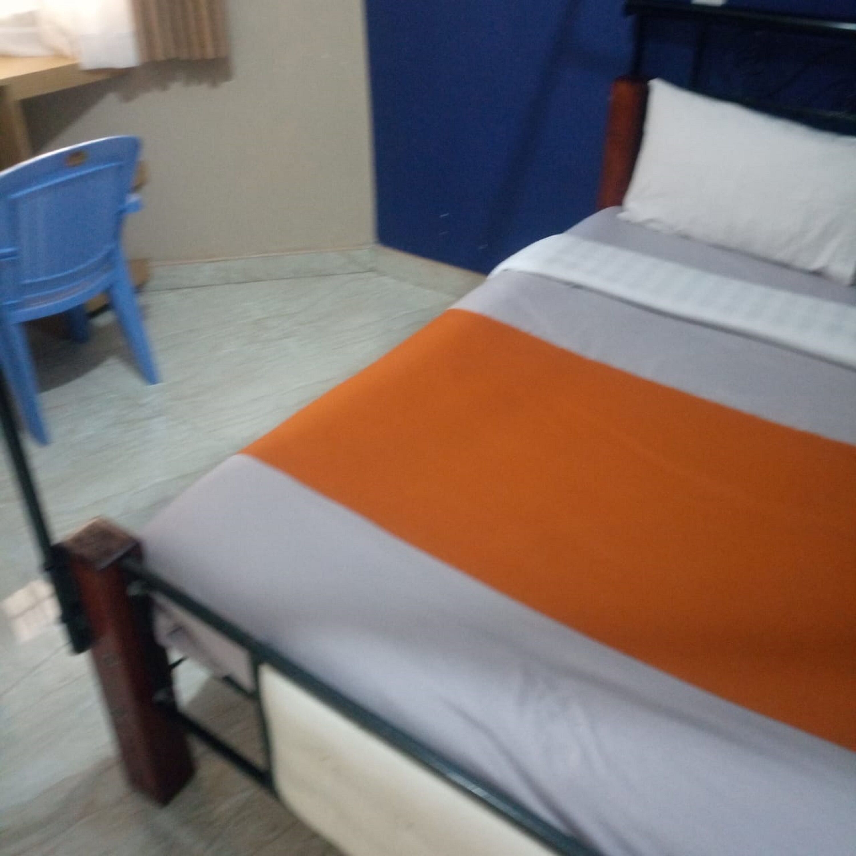 A bed with an orange and grey bedspread, a blue chair, and a wooden desk in a room with a tiled floor.