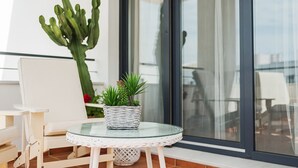 Een balkon met een rieten tafeltje, een witte stoel, een cactus in pot en een glazen bijzettafeltje.
