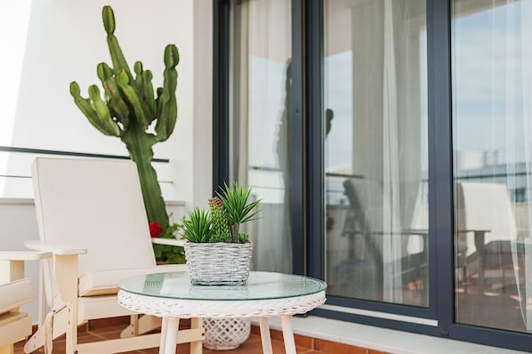 A balcony with a wicker table, a white chair, a potted cactus, and a glass table.