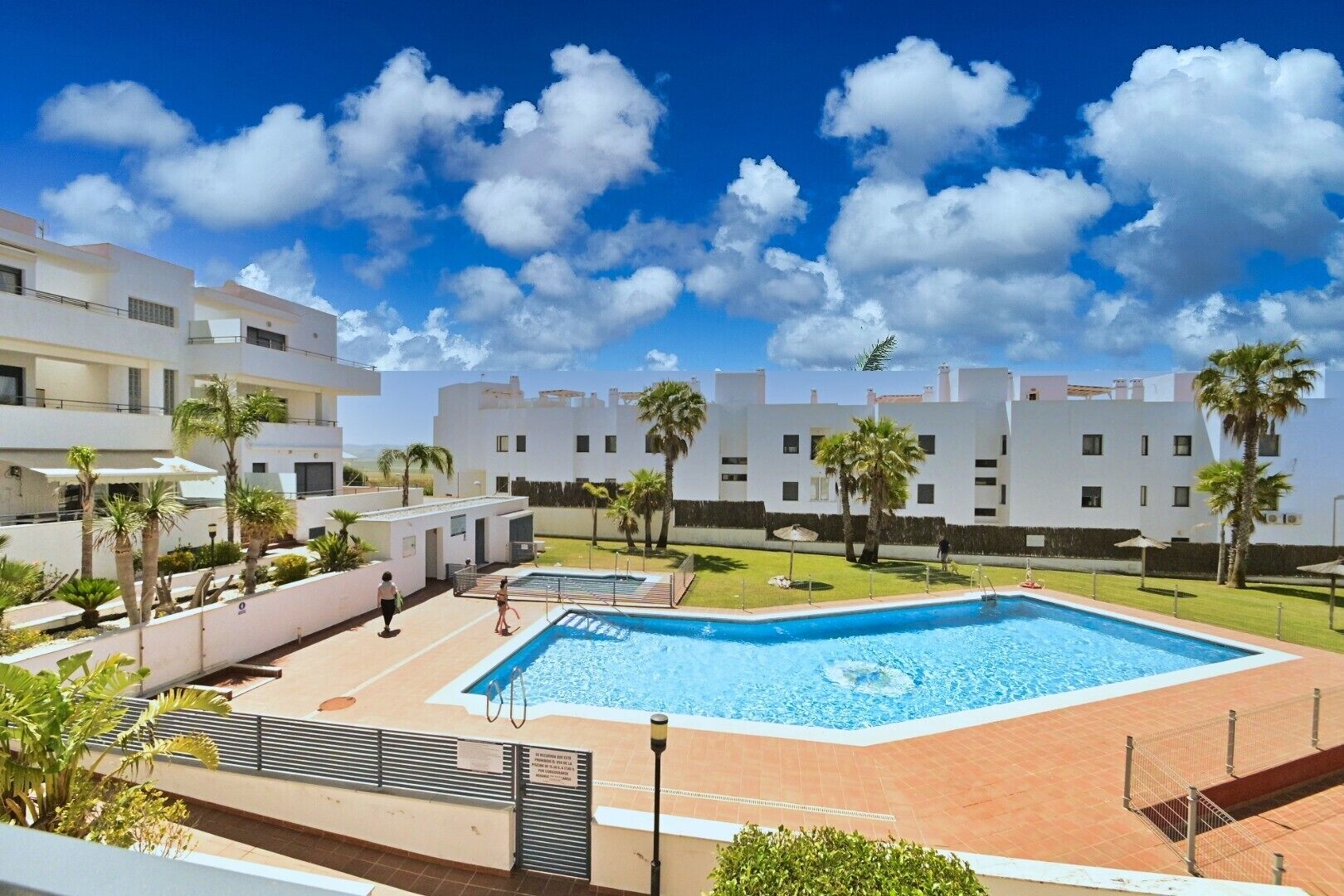 Un ensemble immobilier moderne comprenant une piscine, des palmiers et un ciel d’un bleu éclatant.