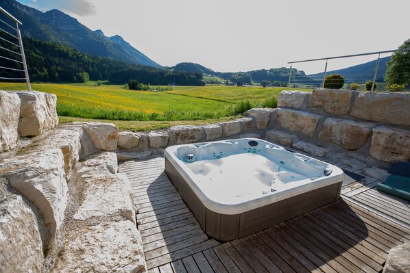 Un jacuzzi en una terraza de madera con paredes de piedra, con vistas a un campo verde y montañas.