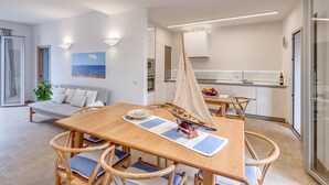 Une cuisine moderne avec des meubles blancs, un coin repas avec une table et des chaises en bois, une maquette de voilier et un tableau représentant un paysage urbain accroché au mur.