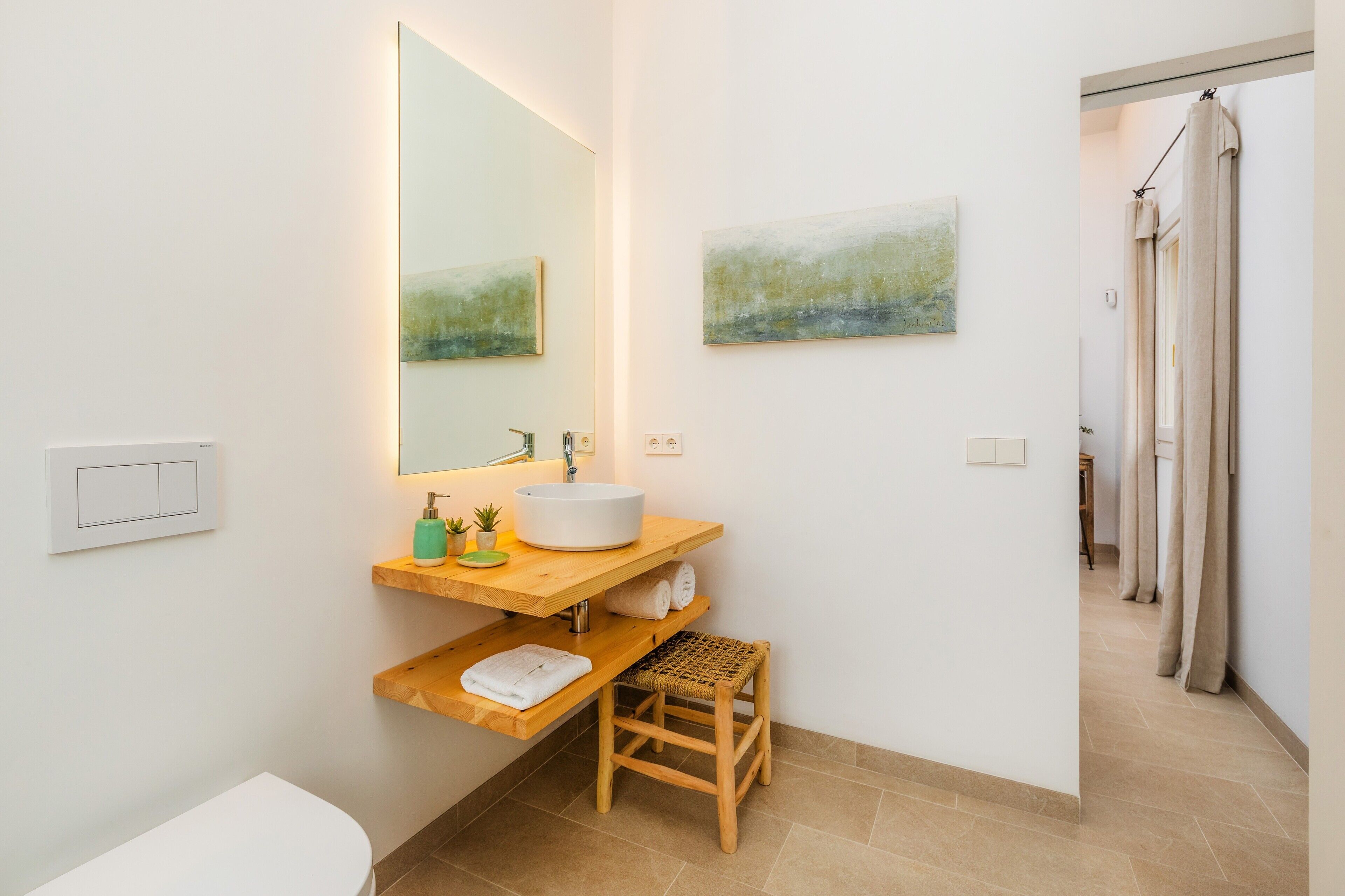 A modern bathroom with a large mirror, a round sink, a wooden shelf, a small stool, and a shower curtain.