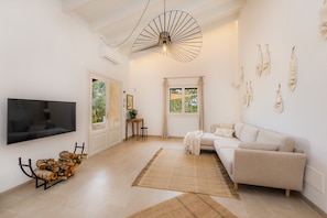 A modern living room with a flat-screen TV, a sofa, a coffee table, a decorative light fixture, and a window with curtains. - Romantic Natural Stone Finca with Pool, Garden, Rooftop Terrace and Wi-Fi (Artá)