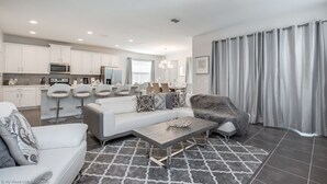 Una sala de estar moderna con sofá, sillón y mesa de centro, una cocina con armarios y electrodomésticos blancos, y un comedor con taburetes de bar.
