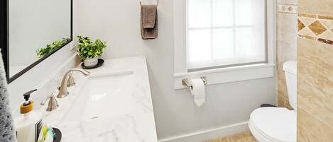A bathroom with a white sink, mirror, and toilet, a window with a towel rack, and a small plant on the counter.