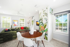 A modern living room with a green sofa, white chairs, a wooden dining table, and a large window with a view.
