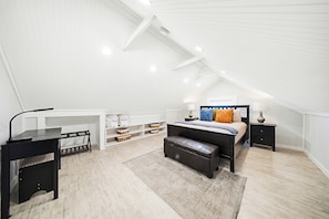 A bedroom with a lofted desk, a bed with pillows, a nightstand, and storage shelves.