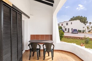 A balcony with a table and chairs, a closed shutter door, and a view of a street with buildings and cars. - Cala Serena Beach 5 — Casa Morrás 43 (Felanitx)