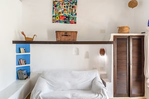 A room with a white sofa, a bookshelf with books, a colorful abstract painting, a wicker basket, and a wooden door. - Cala Serena Beach 5 — Casa Morrás 43 (Felanitx)