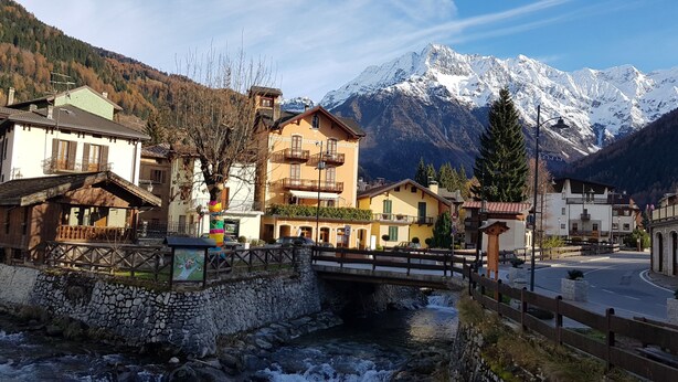 Une ville de montagne avec un pont de pierre enjambant une rivière, des bâtiments dotés de balcons et, en arrière-plan, des montagnes aux sommets enneigés.