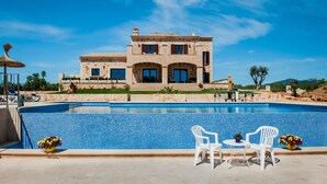 A pool with white chairs and a table, a stone building with arched windows, and a clear blue sky.