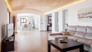 A spacious living area with a sofa, a coffee table, and a TV on a stand. There is a dining area in the background with a table and chairs.