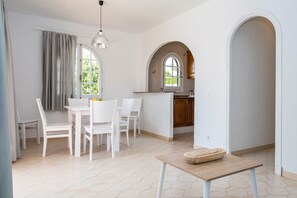 A dining area with a table and chairs, a kitchen with an archway, and a window with curtains.