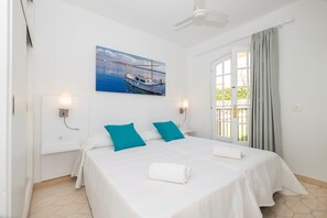 Een tweepersoonsbed met witte en turquoise beddengoed, een plafondventilator, een schilderij van een boot en een raam met gordijnen.