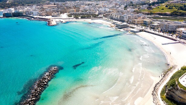 Vista aérea de una ciudad costera con mar azul turquesa, playas arenosas y una zona urbana densa con edificios.