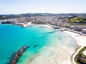 An aerial view of a coastal city with a clear blue sea, sandy beaches, and a dense urban area with buildings.
