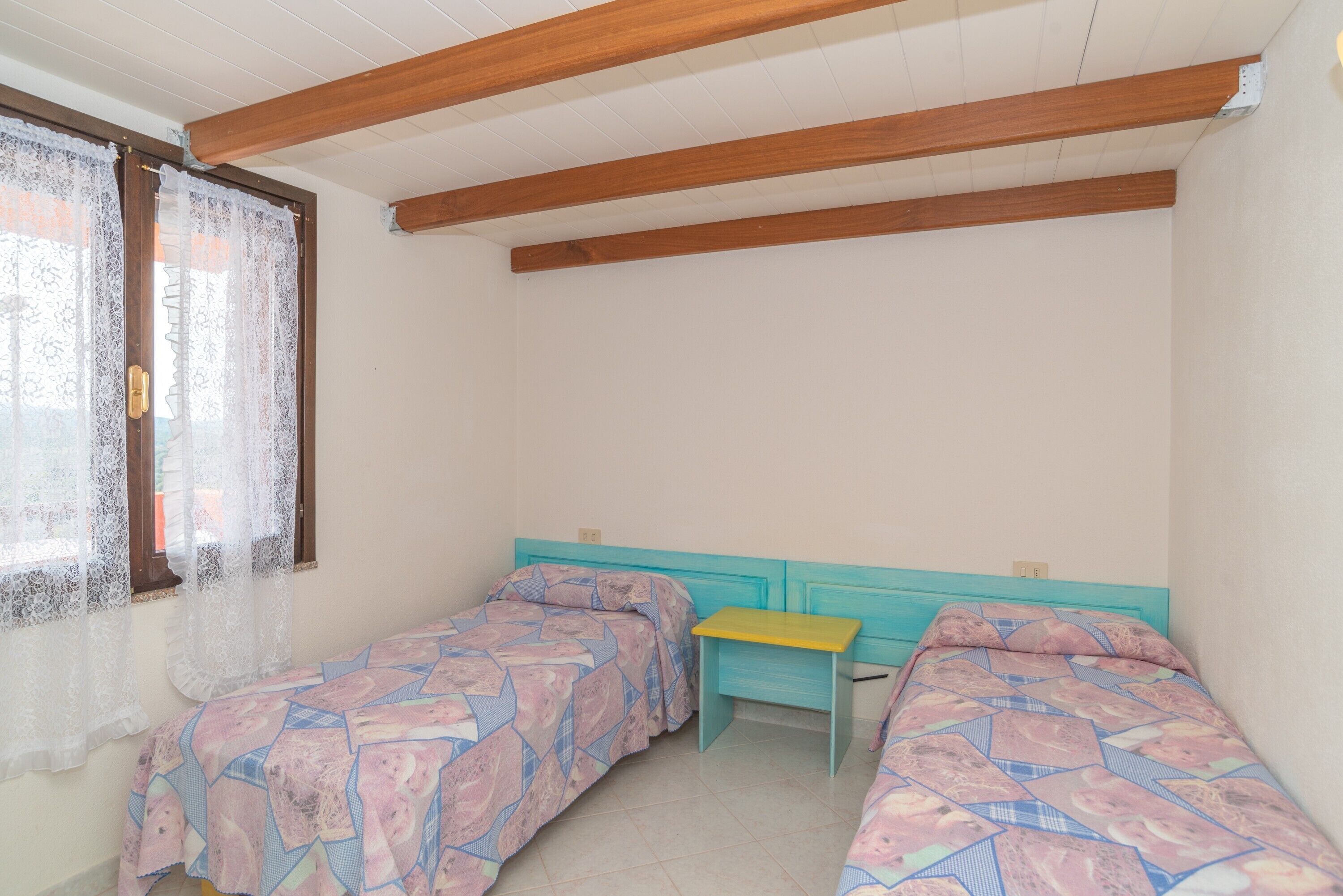 A room with two beds, a small table, and a window with lace curtains.