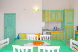 Une pièce avec une table verte, des chaises blanches, une cuisine avec des meubles verts et un dosseret de carrelage bleu et blanc.