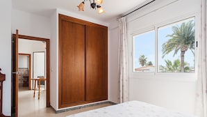 Ein Schlafzimmer mit Bett, einem Holzschrank, einem Fenster mit Vorhängen und Blick auf Palmen.