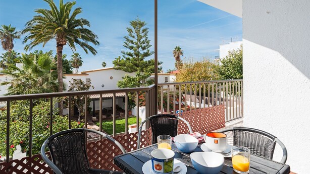 Ein Balkon mit Korbstühlen, einem Tisch mit Frühstücksgebäck und Blick auf Palmen und Gebäude.