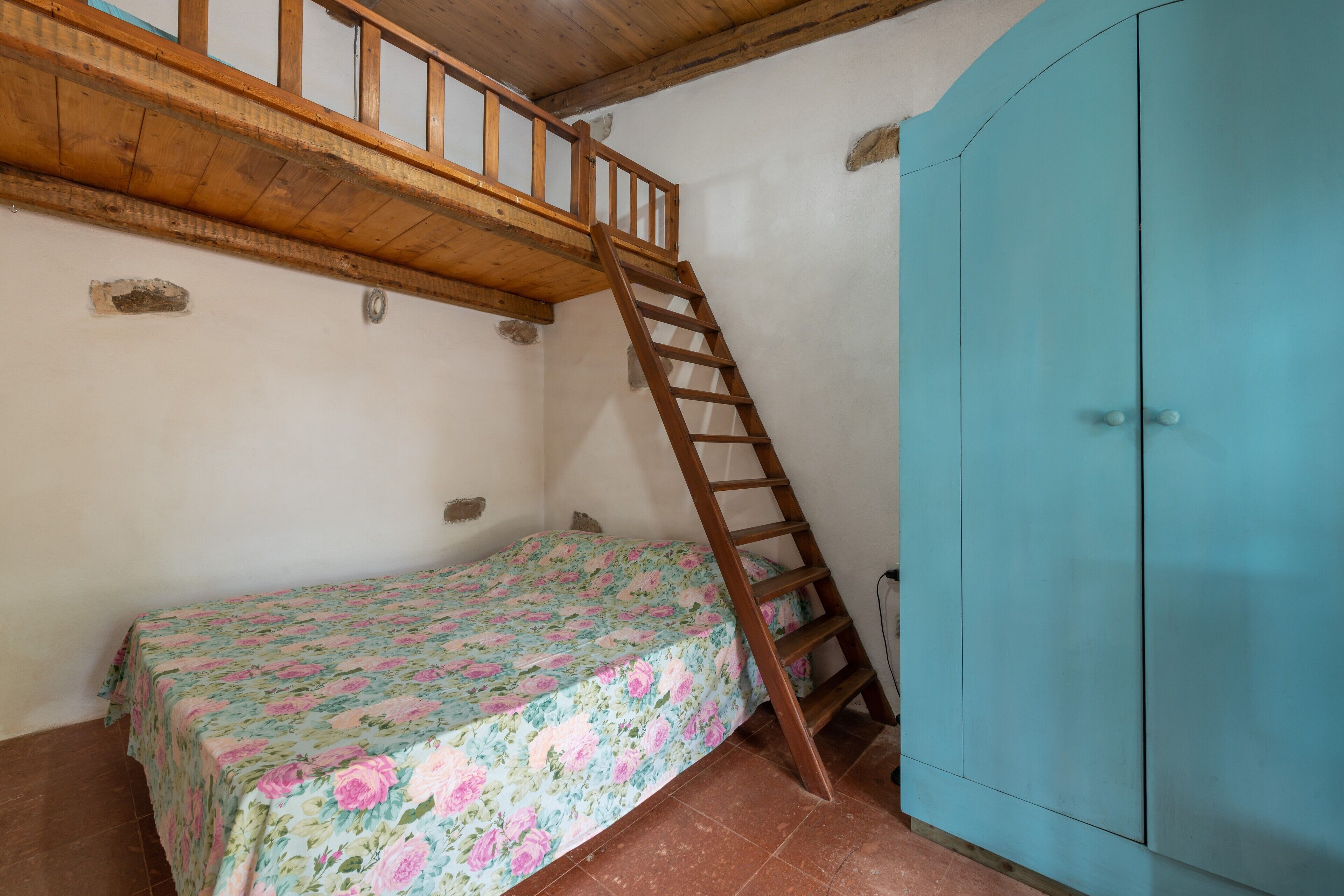 A bedroom with a wooden ladder leading to a loft, a bed with a floral bedspread, and a blue wardrobe.