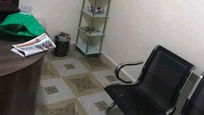 A waiting area with a reception desk, a clock, a trash bin, a green bag, a newspaper, a metal shelf with flowers, and a black chair. - The Loop Guest House and Restaurant (Eldoret)
