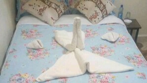 A bed with a floral bedspread and pillows, a water bottle on a nightstand, and a towel folded in the shape of a bird. - The Loop Guest House and Restaurant (Eldoret)