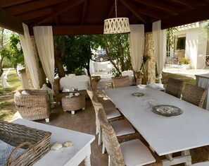 Een terras buiten met rieten meubels, een witte tafel en een hanglamp.