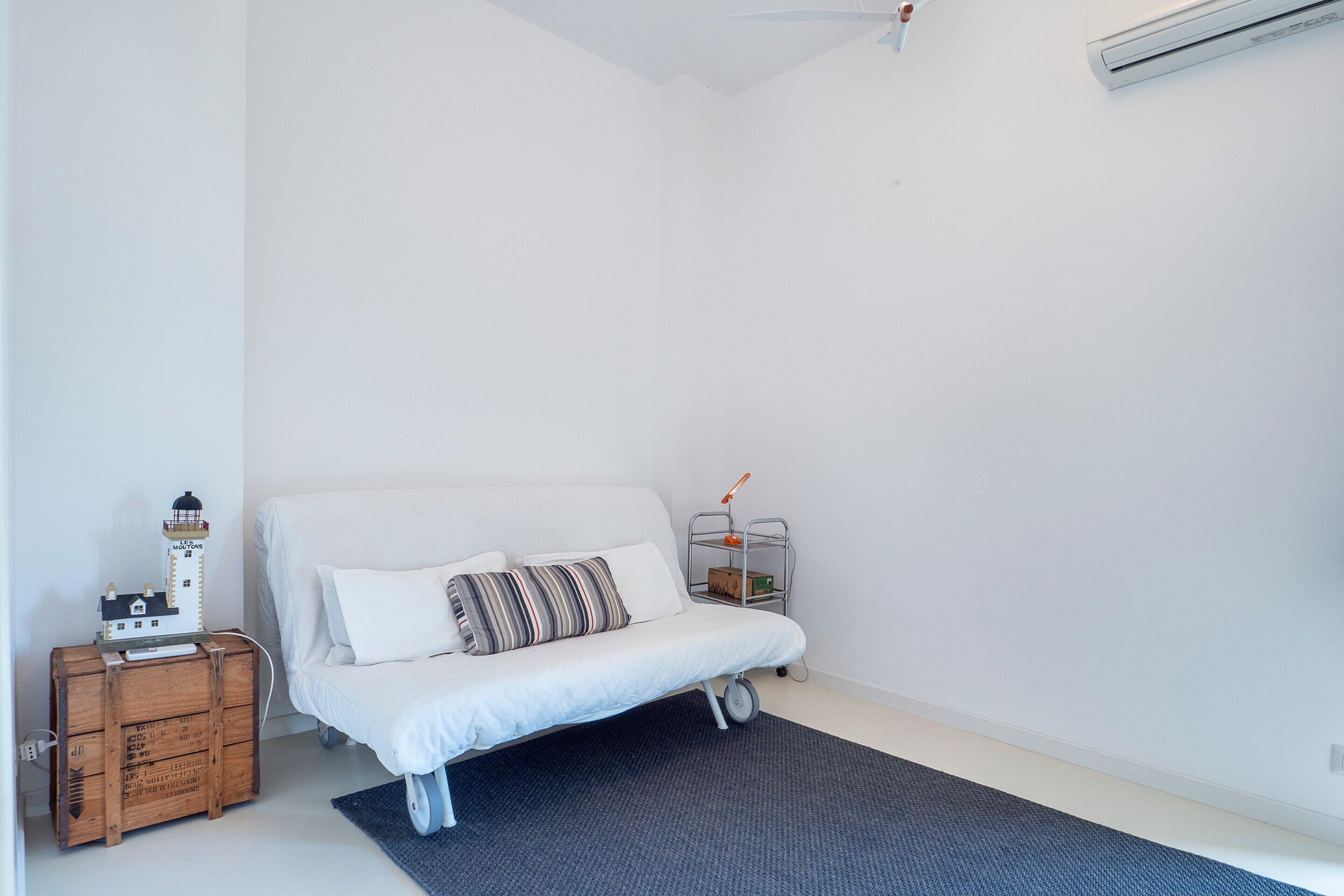 Habitación minimalista con un sofá blanco, un pequeño cofre de madera, una estantería metálica y una maqueta de faro.