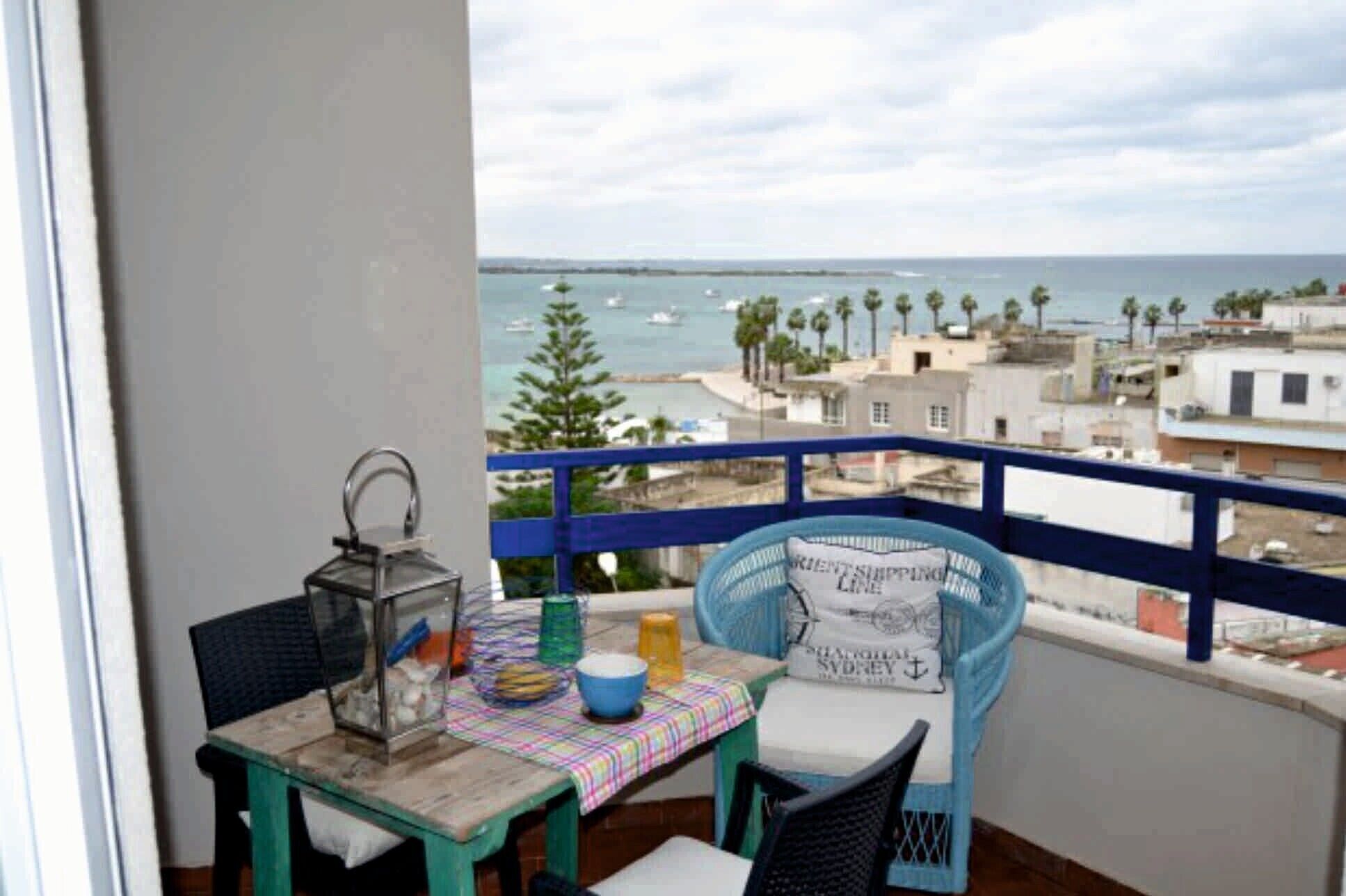 Un balcón con una mesa y sillas, vista al mar y edificios al fondo.