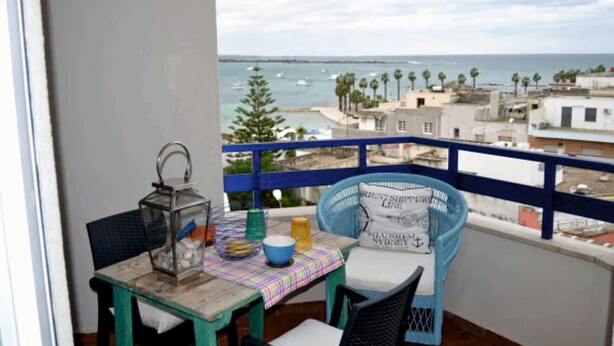 Ein Balkon mit Tisch und Stühlen, Blick auf das Meer und Gebäude im Hintergrund.