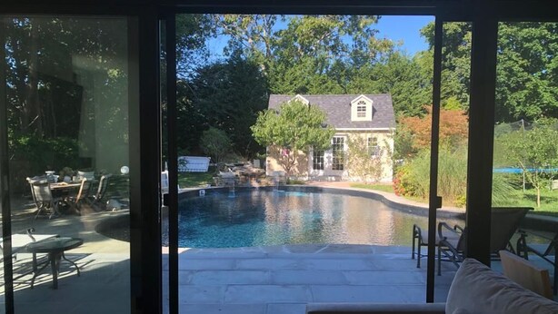 Blick aus einem Zimmer mit Schiebeglastür auf einen Bereich mit Außenpool, eine Terrasse mit Tisch und Stühlen sowie ein Haus im Hintergrund.