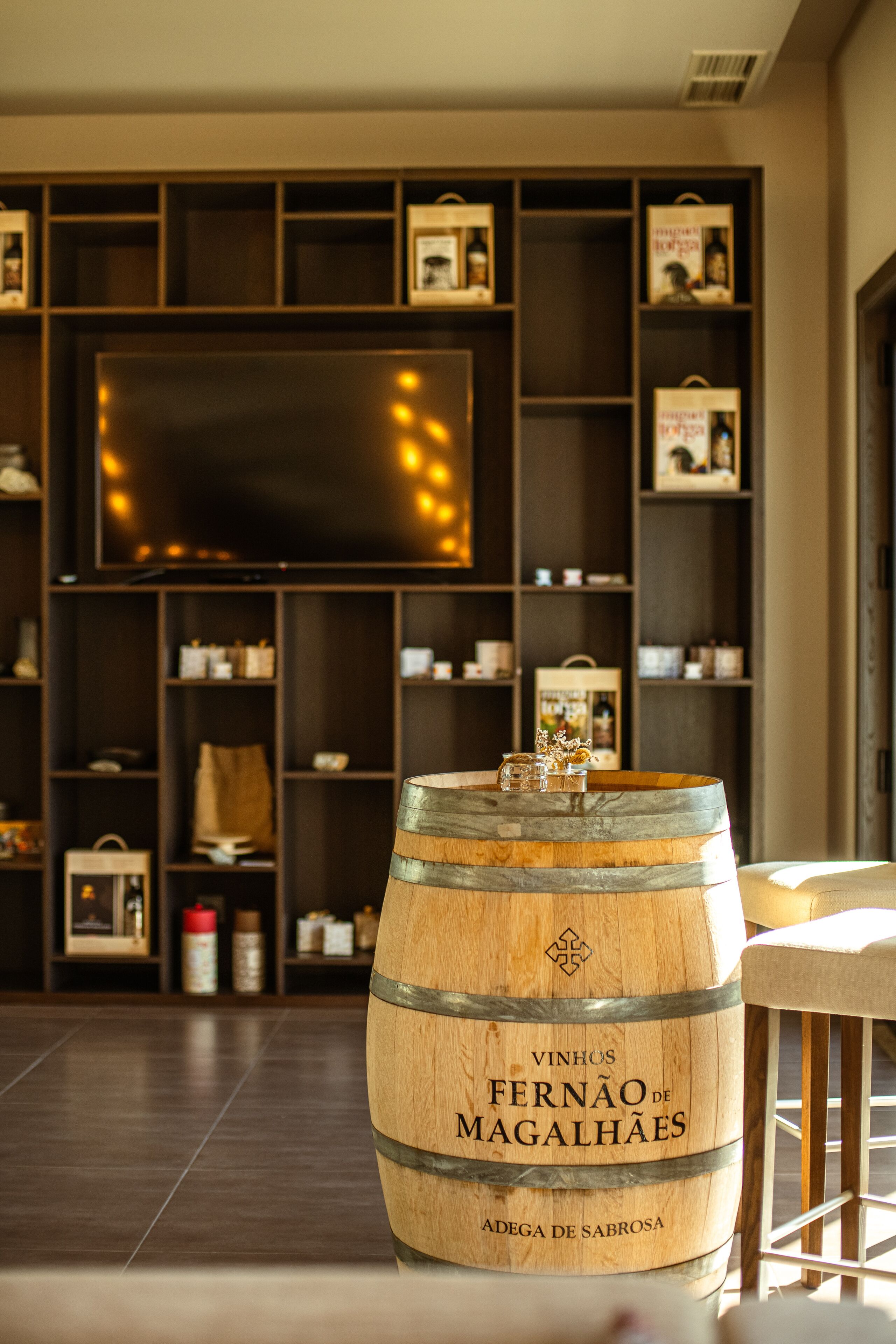 A wooden barrel labeled "Vinhos Fernão de Magalhães" in a room with a large flat-screen TV mounted on the wall and shelves with various items.