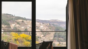 Une pièce dotée d’une grande fenêtre, de deux chaises, d’une petite table et offrant une vue sur un paysage vallonné.