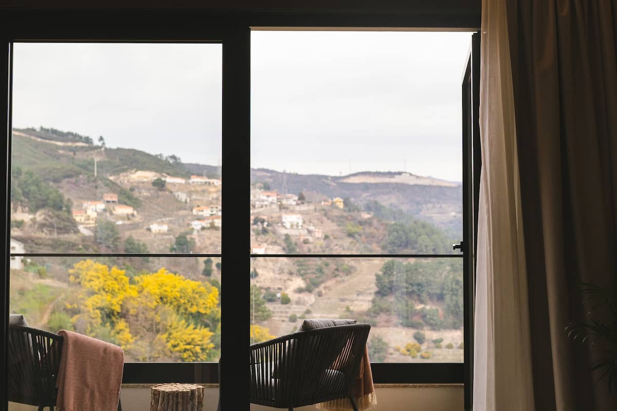 Une pièce dotée d’une grande fenêtre, de deux chaises, d’une petite table et offrant une vue sur un paysage vallonné.