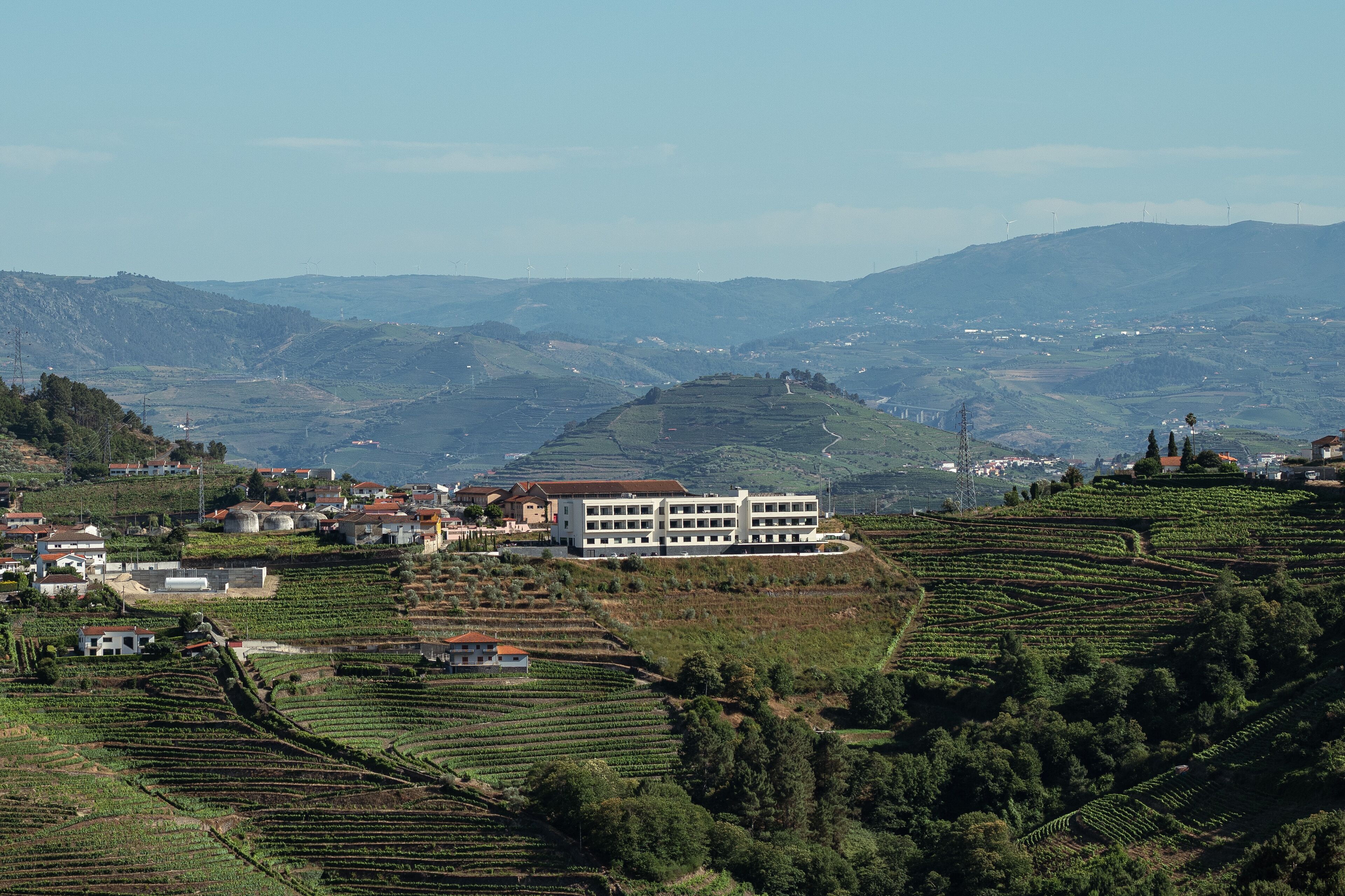 A rural landscape with terraced vineyards, a large white building with multiple floors, and rolling hills in the background.