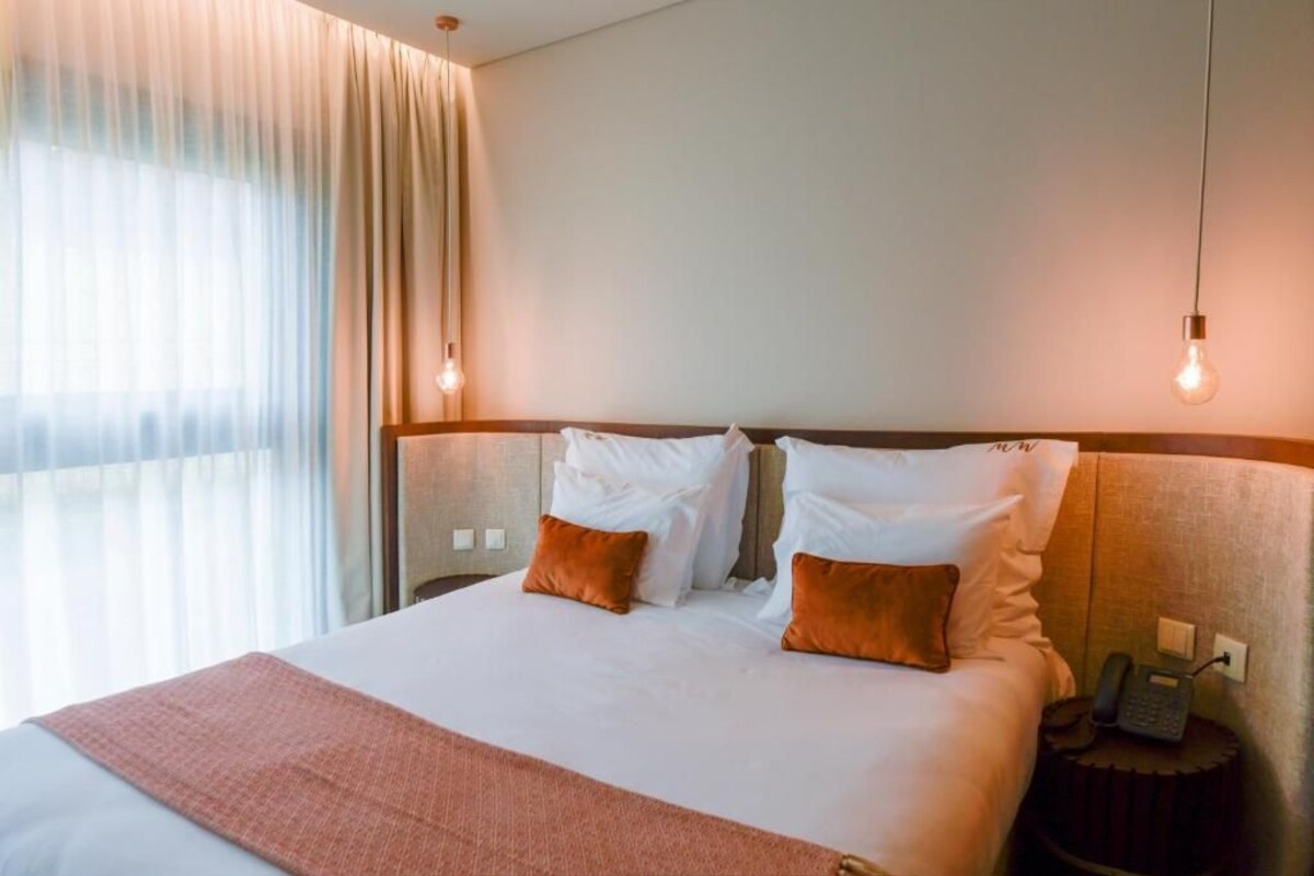 Una habitación de hotel con cama, dos almohadas, una mesita de noche con teléfono y una ventana con cortinas.