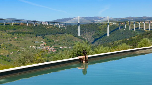 Une personne se détend dans une piscine, avec vue sur un pont et un paysage vallonné.
