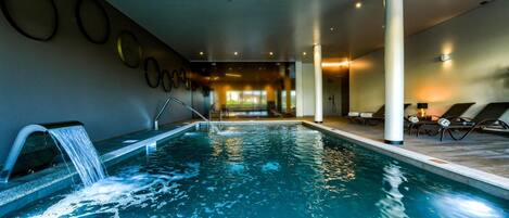 An indoor swimming pool with a waterfall feature, lounge chairs, and a view of the outdoors.