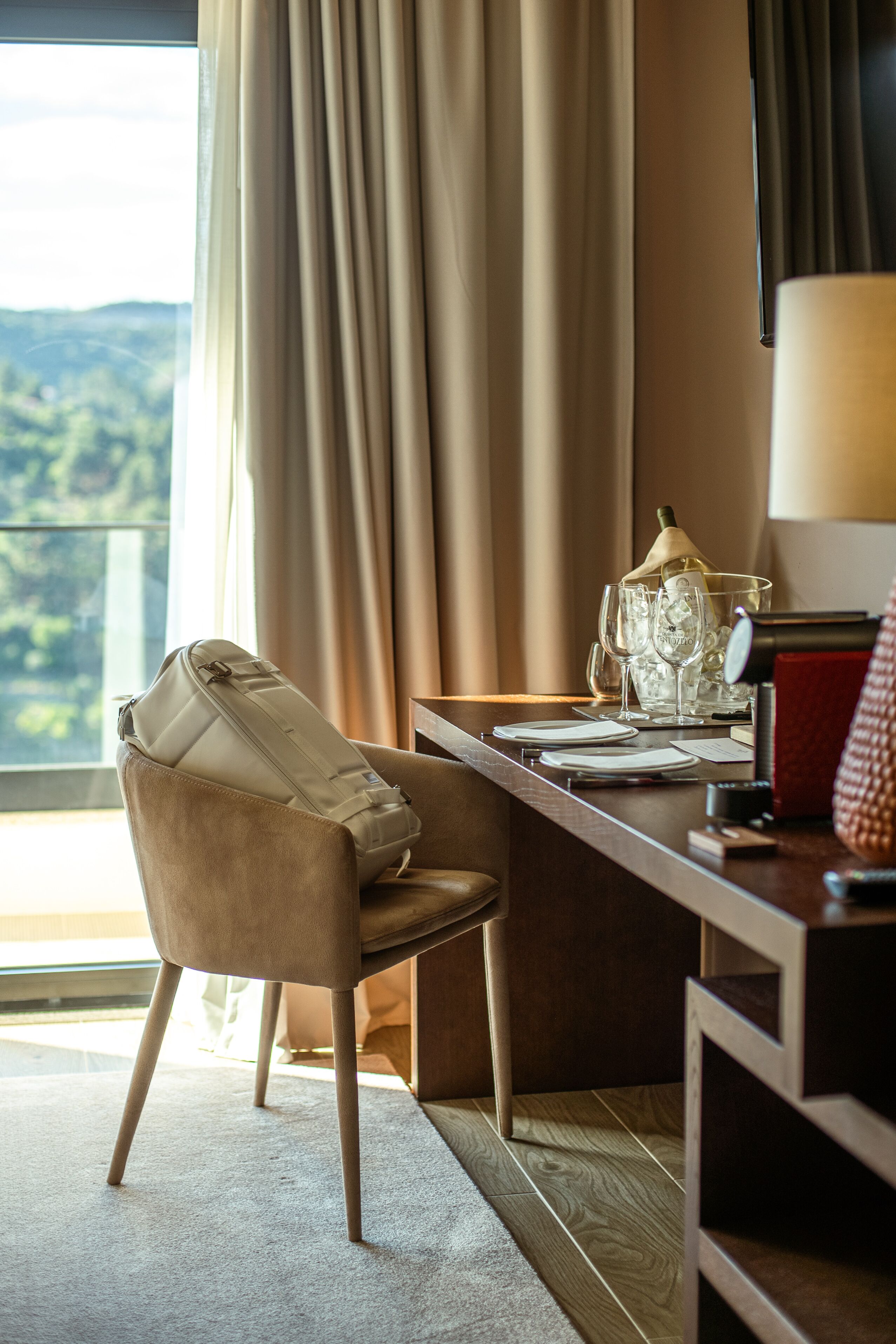A hotel room with a view of the outdoors, a desk with a lamp, and a chair.