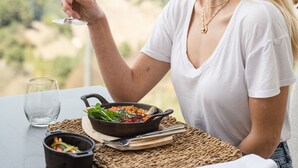 Une personne est assise à une table, devant un repas, avec un verre de vin blanc, et on aperçoit un paysage magnifique en arrière-plan.